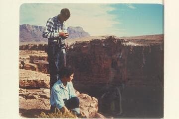 Susie Reilly sits at the edge of Marble Canyon looking down at Badger Creek Rapid.  Pat Reilly prepares for photography