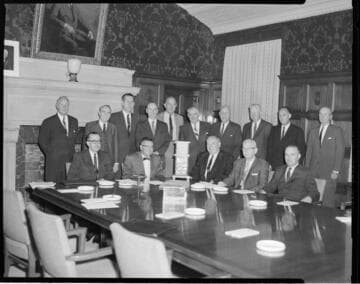 Jack Horton with 4 other executives seated at conference table and 10 additional executives standing behind them in the old General Office board room at 5th & Grand