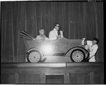 Performance of skit at Edison auditorium.  Man and woman in cardboard car with another man fixing a flat on the car